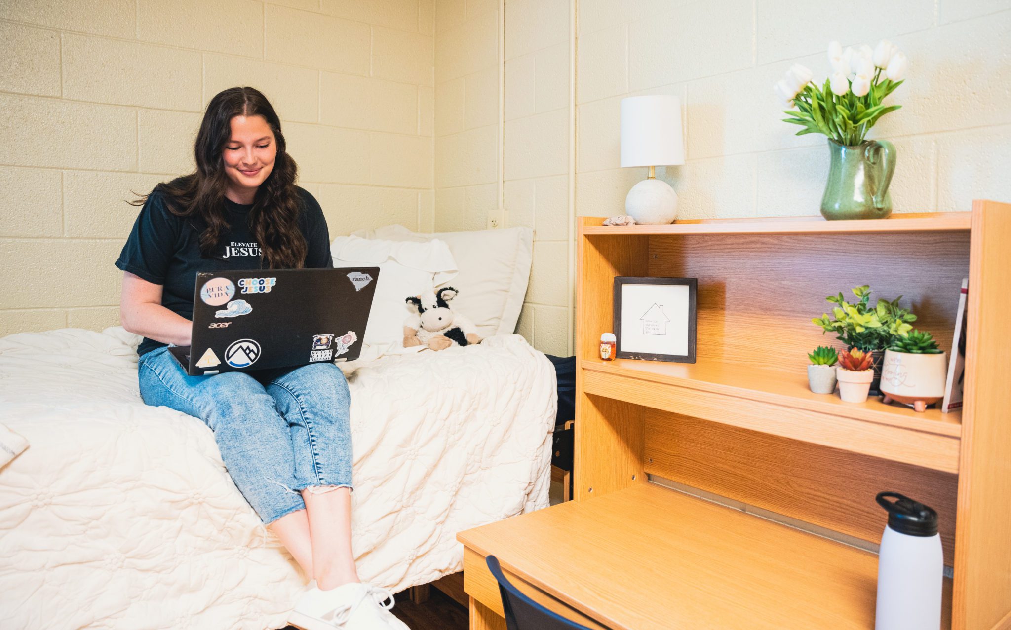 A gril sitting on her bed with her computer in her dorm. There is a desk next to her bed.
