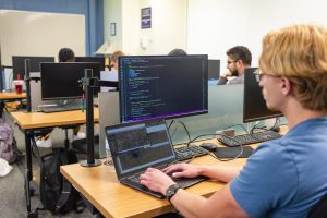 CSU student looking at two computers with code on the screens in a classroom.