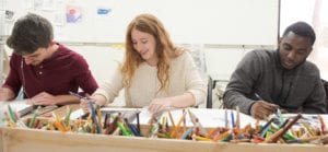 Three art students sketching with pencil on paper with a long wooden box of pencils in front of them
