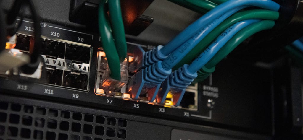 The back of a networking server with many blue and green cables coming out of it.