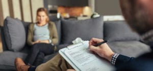 Psychologist taking down notes during therapy session with female patient. Psychotherapist with clipboard and woman patient sitting in background.