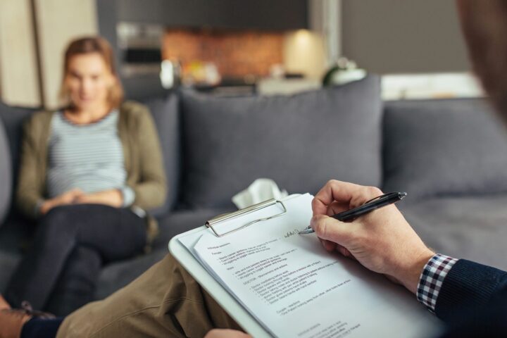 Psychologist taking down notes during therapy session with female patient. Psychotherapist with clipboard and woman patient sitting in background.