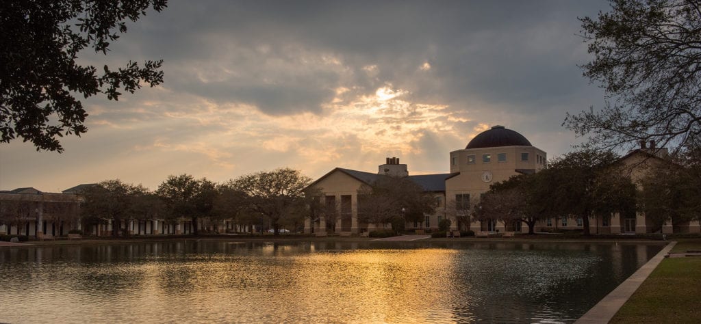 The Science Building with the sun setting behind it casting rays through the clouds. The golden color reflects off of the reflection pond.