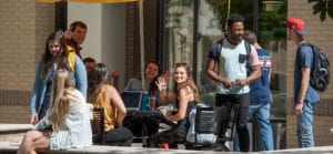 Many students gathered around an outside table in front of the library, some waving at the camera.
