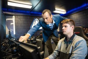 A boy sitting in the racing seat and looking at the screen. There is another man next to him who gives him instructions