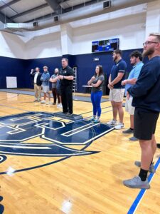 Students standing at the basketball field in the brewer center and listening to a person speaking.
