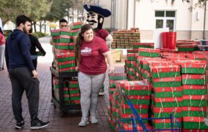 A group of people and Bucky bringing the Christmas boxes all together|Big multiple boxes displayed next to each other to put the filled christmas boxes for children|A party in chapel celebrating the new record for collecting the boxes for children. The University collected 13