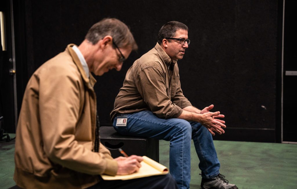 A professor sitting on a box explainig the play called the Holocaust and a man taking notes