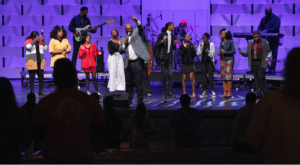 A man speaking in chapel with a singing band behind him
