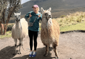 A woman is between two alpaca's with a beautifull view behind her